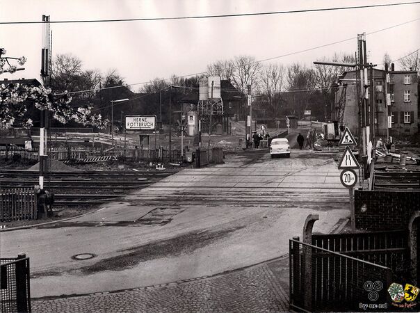 Bild 1: Blick vom Stellwerk Hrb des Bahnhofs Herne-Rottbruch nach Nordosten. Ein Personenzug Richtung Bochum steht auf der neuen Brücke (zugleich Haltepunkt Herne-Rottbruch), die später über die Autobahn (heute A 43) führen wird. Die Ausschachtungsarbeiten für die Autobahn, die hier bekanntlich in einem Einschnitt verläuft, sind in vollem Gange. Im Vordergrund der Bahnübergang Rottbruchstraße, die Straße mündet rechts in den Grenzweg. Die Behelfsbrücke rechts steht ebenfalls im Zusammenhang mit den Ausschachtungsarbeiten.