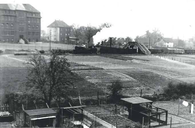 Am linken Bildrand die Vellwigschule. In der Bildmitte ist der Fußgängerübergang über die Hafenanschlußbahn, von der Zeche Mont-Cenis zum Hafen Friedrich der Große III/IV, zu sehen, welche auch von Zügen der Zeche Constantin benutzt wurde. Einer dieser Züge mit einer Lok Henschel Typ Frankfurt, wohl Constantin Nr. 14 oder 16, mit einem Zug Kübelwagen, unterquert die Fußgängerbrücke. Dort befindet sich heute die Fußgängerbedarfsampel an der Sodinger Straße. Im Vordergrund sind Gärten der Teutoburgiasiedlung, im Hintergrund weitere Häuser des Dorfes Börnig zu erkennen.